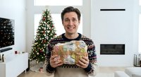 Happy Caucasian man in festive sweater holding wrapped Christmas gift in modern living room with decorated tree and fireplace during holiday season.