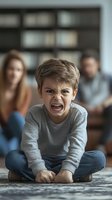 Young boy showing intense anger while sitting cross-legged on floor, wearing gray sweater and jeans, with parents blurred in background during family therapy session.