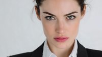 Professional young Caucasian businesswoman with intense green eyes and pink lips in black suit jacket looking directly at camera against neutral background.