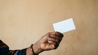 Adult male hand with dark skin tone holding blank white card against neutral beige background for business, identity, or promotional messaging.