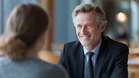 Mature Caucasian businessman in dark suit smiling warmly during office meeting with colleague, professional corporate environment with blurred background.
