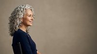 Mature woman with long gray curly hair, Caucasian ethnicity, wearing dark navy sweater, smiling in profile against neutral beige background for portrait and lifestyle use.