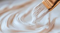 Artistic close-up of white paint swirling from paintbrush bristles creating smooth circular patterns against neutral background, showing fluid creative motion.