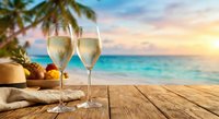 Two champagne glasses on wooden deck overlooking tropical beach at sunset with palm trees and turquoise ocean for vacation celebration and romantic getaway.
