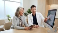 Male doctor consulting with mature female patient using computer screen in modern medical office setting for healthcare discussion.