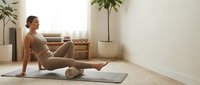 Woman with reddish-blonde hair, Caucasian ethnicity, performing self-massage on leg muscles using foam roller during home fitness routine in minimalist bedroom.