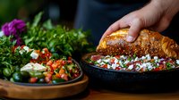 Fresh Mediterranean salad with crumbled feta cheese, tomatoes and herbs served in rustic bowls, hand placing artisanal bread, moody restaurant setting.
