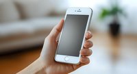 Female hand holding white smartphone with blank screen against blurred living room background with houseplant and sofa, mockup for app presentation.