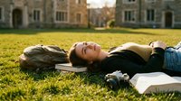 Young woman lying on grass reading book in university courtyard during golden hour sunset with historic stone buildings in background for education lifestyle content.