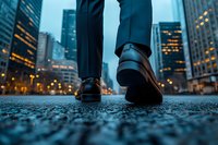 Close-up view of businessman's feet in leather shoes walking on urban street at dusk, with illuminated skyscrapers and city lights creating atmospheric mood.