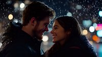 Young couple sharing intimate moment in falling snow with city lights bokeh background creating romantic winter evening atmosphere.