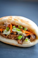 Asian steamed bun filled with tender braised pork belly, topped with fresh carrots, green onions, and shredded cabbage on dark background. Perfect for food menu.