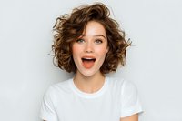 Excited young woman with curly brown hair expressing joy and surprise in white t-shirt against light background for lifestyle and emotion concepts.