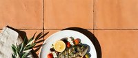 Grilled fish fillet with lemon, tomatoes, and herbs on white plate against terracotta tile background, overhead flat lay for culinary and food styling use.