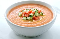Refreshing cold gazpacho soup garnished with diced cucumber and tomatoes in white ceramic bowl on light background, traditional Spanish cuisine.