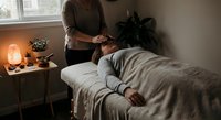 Professional massage therapist performing relaxing treatment on client in cozy spa room with warm lighting and plants creating peaceful wellness atmosphere.