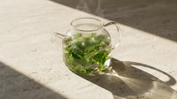 Fresh mint tea in clear glass teapot with green leaves steeping in hot water on marble surface with natural sunlight shadows for wellness and relaxation.