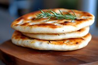 Fresh homemade naan bread stack garnished with rosemary sprigs on wooden cutting board, golden-brown charred spots, soft fluffy texture, close-up food photography.