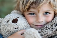 Young blonde child with blue eyes smiling while hugging soft teddy bear outdoors. Cozy childhood moment capturing innocence and comfort for family content.