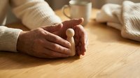 Elderly hands gently holding white pill or medication on wooden table with coffee cup, representing healthcare, medicine management, and senior wellness.