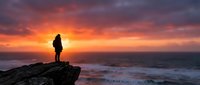 Silhouette of a person standing on rocky cliff overlooking ocean at dramatic sunset with golden and orange sky, waves crashing below, adventure and contemplation theme.