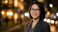 Professional Asian businesswoman with glasses smiling confidently in modern office environment with warm bokeh lighting for corporate presentations.