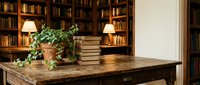 Wooden desk with stacked books and potted plant in front of illuminated bookshelf with warm lighting, suitable for home office and library interior design projects.