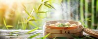 Asian bamboo steamer with dim sum dumplings on wooden table against green bamboo leaves background with steam rising and soft natural light.