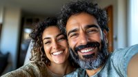 Middle Eastern couple taking selfie at home, showing genuine happiness with bright smiles. Man with gray beard and woman with curly hair lean close together in casual setting.