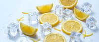 Fresh lemon slices, wedges, zest curls, and melting ice cubes scattered on a white wet surface, evoking summer refreshment.