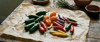 Colorful fresh chili peppers variety on parchment paper with herbs and spices for cooking and culinary preparation on rustic wooden table.