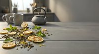 Dried lemon slices, green tea leaves, and fresh mint scattered on a rustic wooden table with a teapot and mugs in the background.