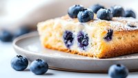 Fresh blueberry cake slice dusted with powdered sugar on gray ceramic plate, scattered berries around, extreme close up macro food photography.