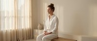 Young Asian woman in white pajamas sitting peacefully on bed in bright bedroom with natural sunlight streaming through curtains creating serene morning atmosphere.