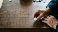 Senior male hand with age spots resting on wooden Go board during game, wearing dark blue sleeve, strategic thinking and traditional Asian board game concept.