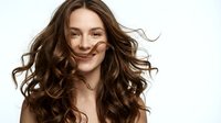 Young Caucasian woman with long curly brown hair in motion, smiling joyfully with natural makeup against white background for beauty and haircare campaigns.