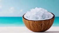 Coconut shell bowl filled with white sea salt crystals on tropical beach with turquoise ocean background for spa wellness and natural skincare products.