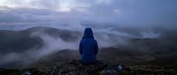Person in blue jacket sitting on rocky mountain peak overlooking misty valleys and distant hills at dusk, contemplative outdoor adventure scene.