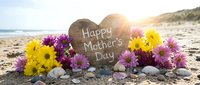 Heart-shaped stone with Happy Mother's Day message surrounded by colorful flowers on sandy beach at sunset for greeting cards and celebrations.