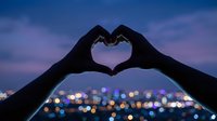 Hands forming heart shape silhouette against twilight city skyline with bokeh lights. Romantic gesture expressing love and connection during blue hour sunset.