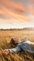 Young Caucasian woman lying in golden field grass at sunset, arms outstretched, eyes closed, enjoying freedom and peaceful relaxation outdoors.
