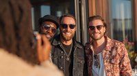Three diverse young men in sunglasses smiling at camera during golden hour photoshoot. Casual trendy fashion lifestyle portrait for social media content.