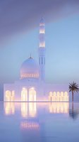 Illuminated white mosque with tall minaret reflecting in water at dusk, soft purple and blue sky creates ethereal atmosphere with single palm tree silhouette.