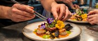 Chef plating gourmet dish with tweezers, arranging colorful edible flowers and vegetables on white plate for fine dining restaurant presentation.