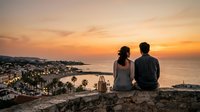 Couple sitting on rocky cliff overlooking coastal town and ocean at sunset, enjoying romantic evening view together.