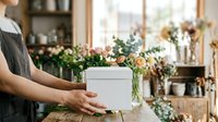 White gift box held by florist in apron at flower shop counter with fresh blooms and greenery arrangements displayed in background for retail and small business use.