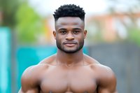 Young athletic African American man with muscular build and short dark hair smiling confidently at camera, shirtless upper body portrait against blurred outdoor background.