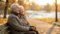 Happy senior couple sitting together on park bench during golden hour autumn sunset enjoying peaceful retirement moment.