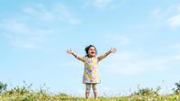Young child with dark hair and light skin tone wearing a colorful floral shirt, standing in open field with arms raised in joy and freedom against blue sky.