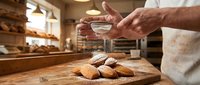 Professional baker dusting powdered sugar on fresh madeleines in warm bakery kitchen with golden lighting and wooden surfaces.